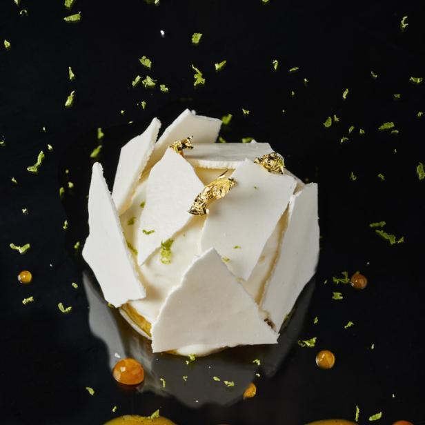 Ein Dessert mit weißer Mousse, Baiserstücken, Blattgold und einer gelben Sauce auf einem schwarzen Teller.
