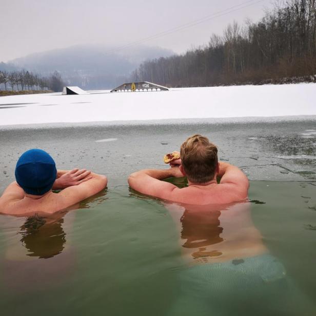 Zwei Männer baden in einem eisigen See und einer isst ein Gebäck.