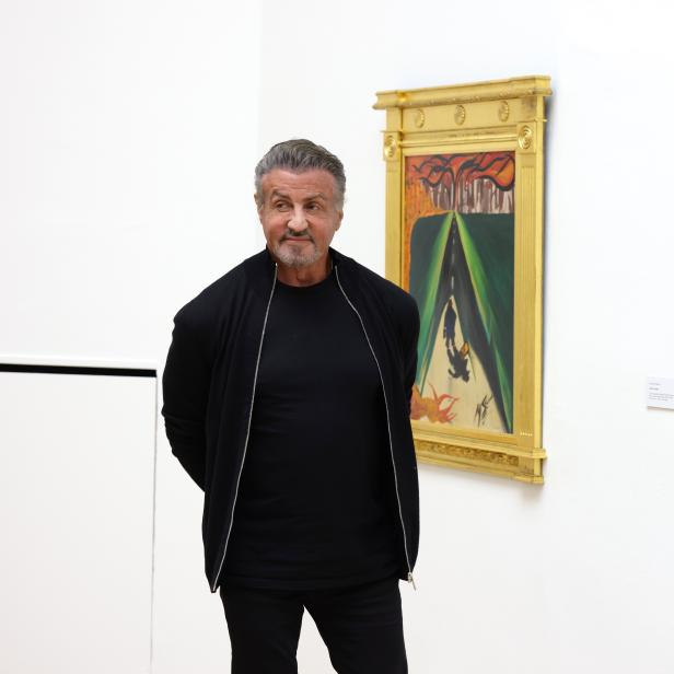 Sylvester Stallone steht vor zwei gerahmten Gemälden in einer Kunstgalerie.