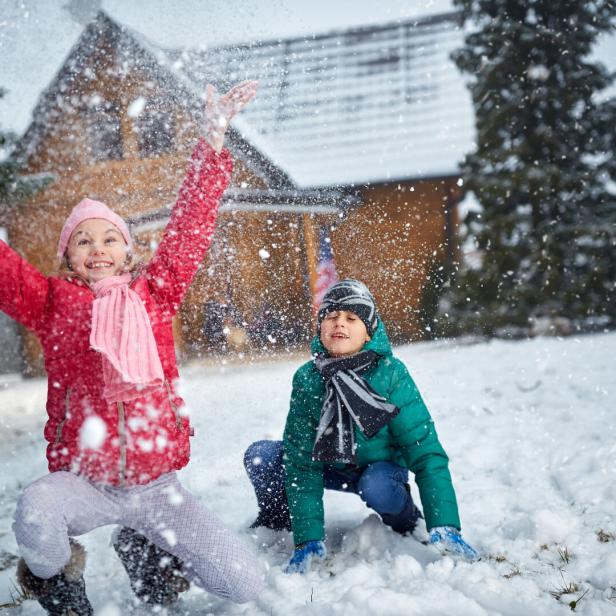Zwei Kinder spielen lachend im Schnee vor einem Haus, während Schneeflocken um sie herum wirbeln.