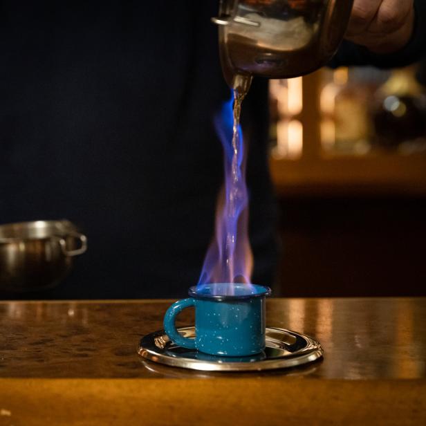 Ein Barkeeper gießt eine brennende Flüssigkeit in eine blaue Tasse.