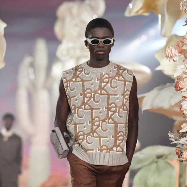 Ein dunkelhäutiges Model mit Sonnenbrille und Strickweste auf einem Laufsteg.