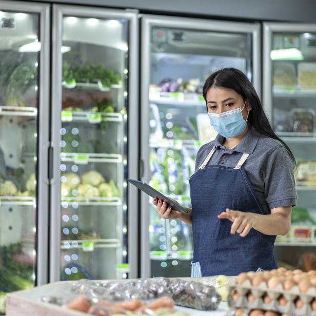 Eine Verkäuferin mit Maske überprüft die Waren in einem Supermarkt mit einem Tablet.