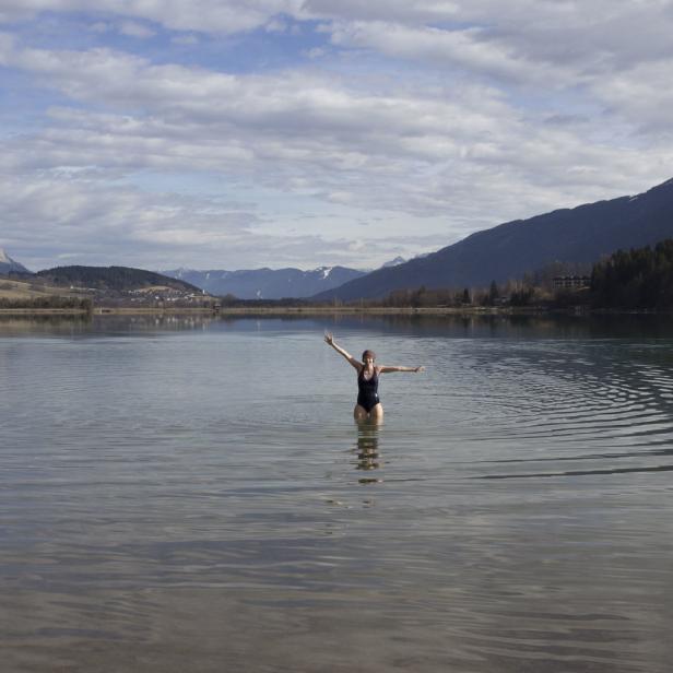 Eine Frau steht mit ausgestreckten Armen in einem See vor einer Bergkulisse.