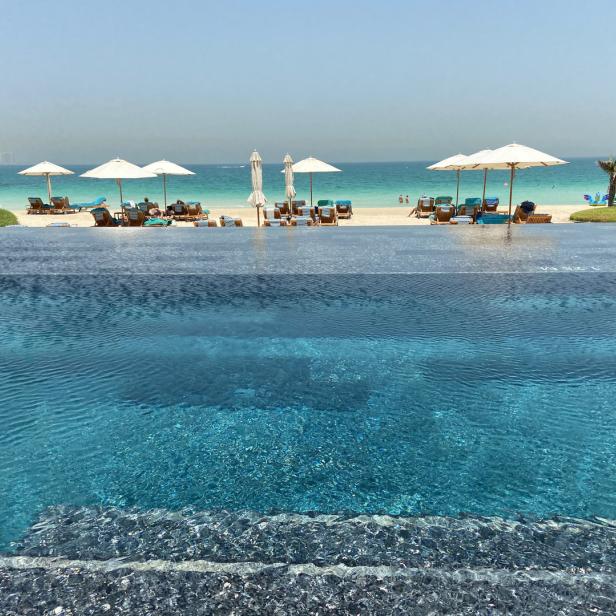 Blick von einem Pool auf einen Strand mit Sonnenschirmen und Liegen.