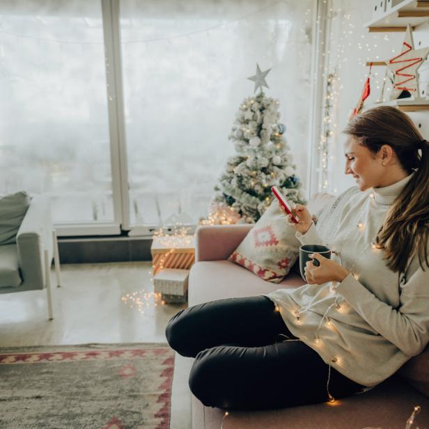 Eine Frau sitzt mit einem Getränk auf dem Sofa und schaut auf ihr Handy.