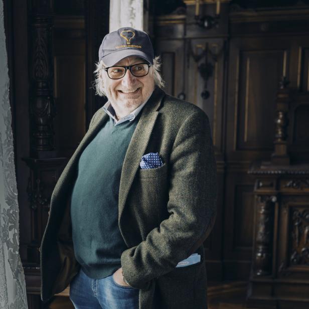 Ein lächelnder Mann mit Brille und Kappe posiert in einem eleganten Raum.