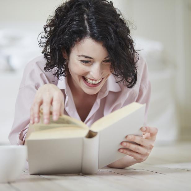 Eine lachende Frau liest ein Buch, während eine Tasse neben ihr auf dem Boden steht.