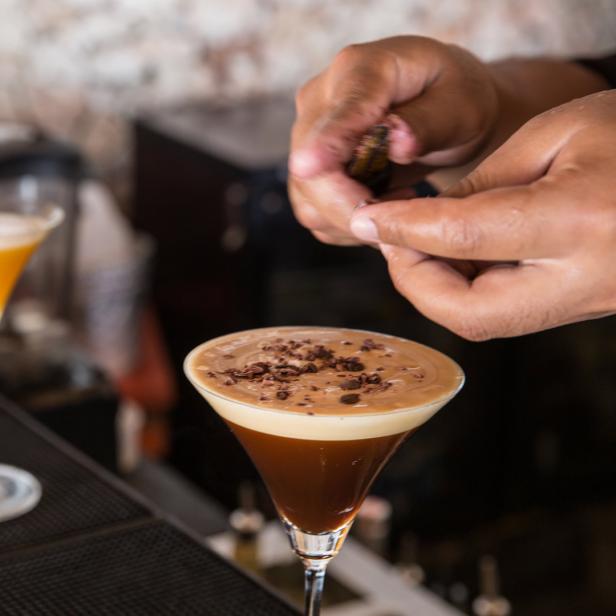 Ein Barkeeper garniert einen Espresso Martini mit Schokoladenraspeln. Im Hintergrund weitere Cocktails.