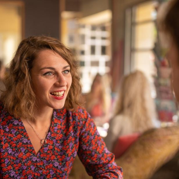 Eine lächelnde Frau mit blumigem Oberteil sitzt in einem Café und unterhält sich.