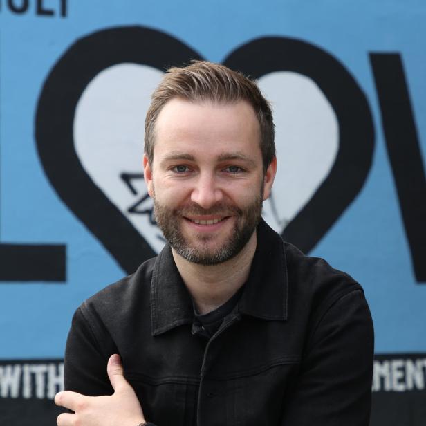 Ein Mann mit Bart lächelt vor einer Wand mit einem Herz-Graffiti.