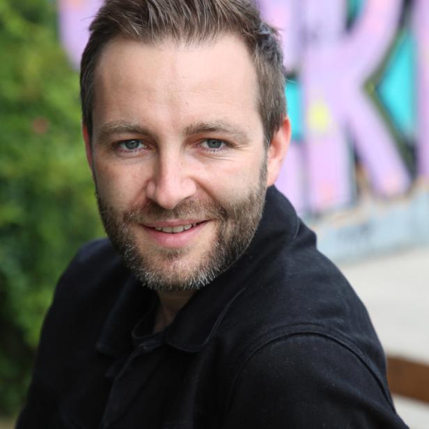 Ein lächelnder Mann mit Bart und dunkler Jacke vor einer Graffiti-Wand.