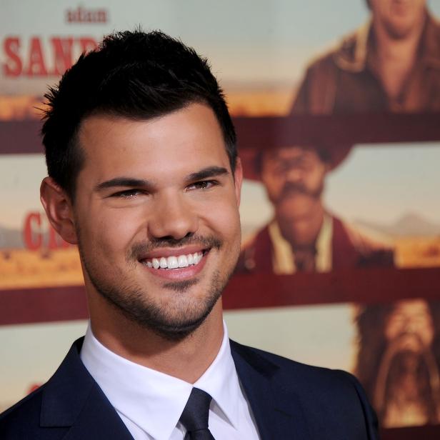 Taylor Lautner lächelt vor einem Poster für den Film „The Ridiculous 6“.