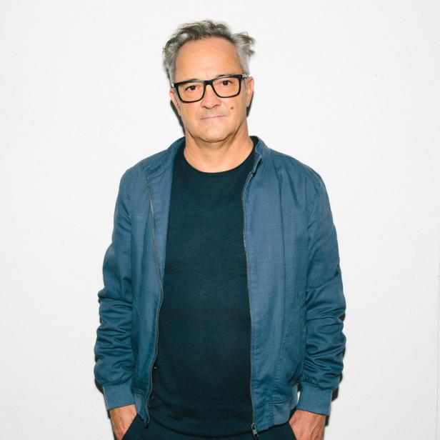 Ein Mann mit Brille und blauer Jacke steht vor einer weißen Wand.