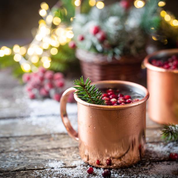 Zwei Kupferbecher mit Getränken und Preiselbeeren auf einem Holztisch mit weihnachtlicher Dekoration.