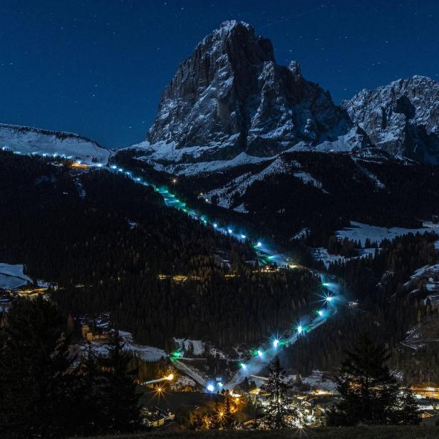 Eine Skipiste schlängelt sich nachts beleuchtet einen Berg hinab.