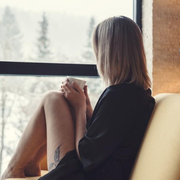 Eine Frau sitzt mit einer Tasse auf einem Sofa und blickt aus dem Fenster auf verschneite Bäume.