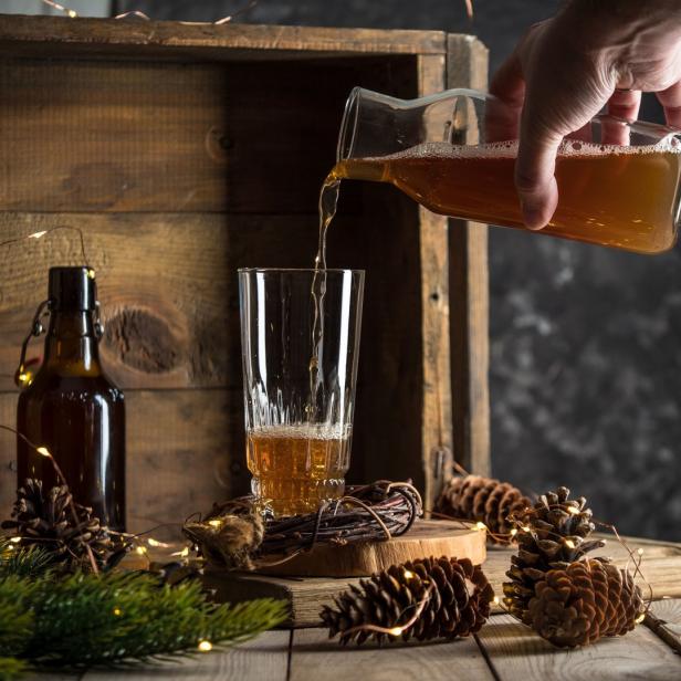 Ein Getränk wird in ein Glas gegossen, umgeben von Tannenzapfen und Lichtern.