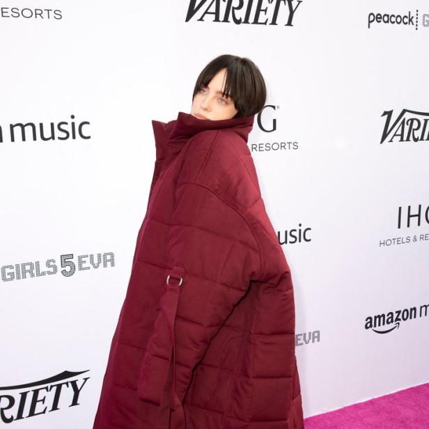Billie Eilish trägt einen übergroßen, burgunderroten Daunenmantel auf einem pinkfarbenen Teppich.