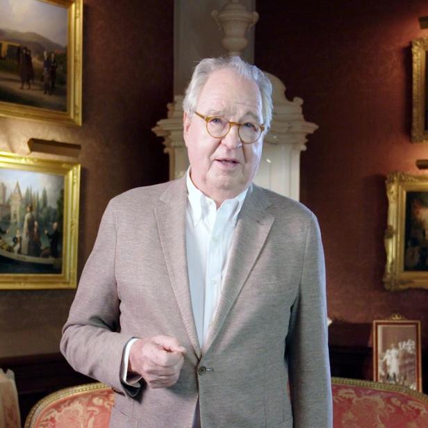 Ein Mann mit Brille und hellem Sakko steht in einem Raum mit Gemälden.