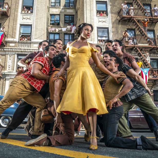 Eine Frau in einem gelben Kleid steht inmitten einer Gruppe von Tänzern vor einem Backsteingebäude mit puertoricanischen Flaggen.