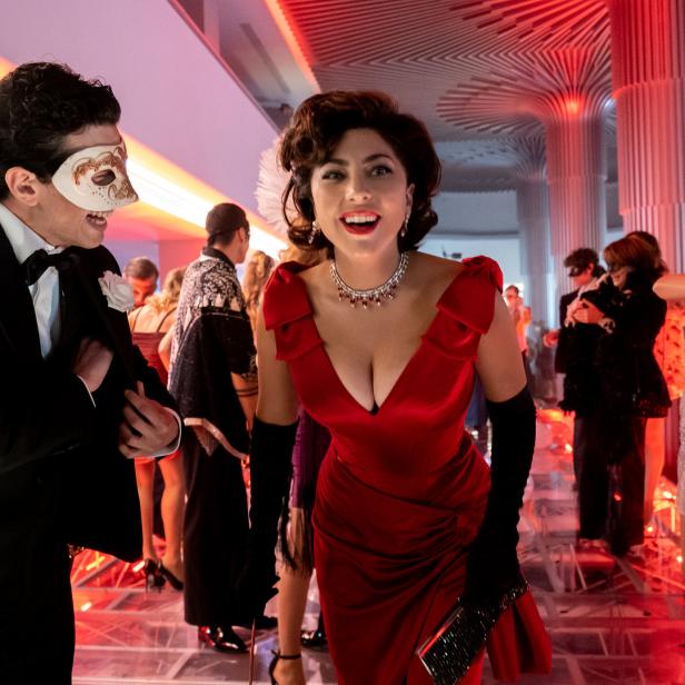 Lady Gaga, in einem roten Kleid, lächelt auf einer Maskerade mit anderen Gästen.