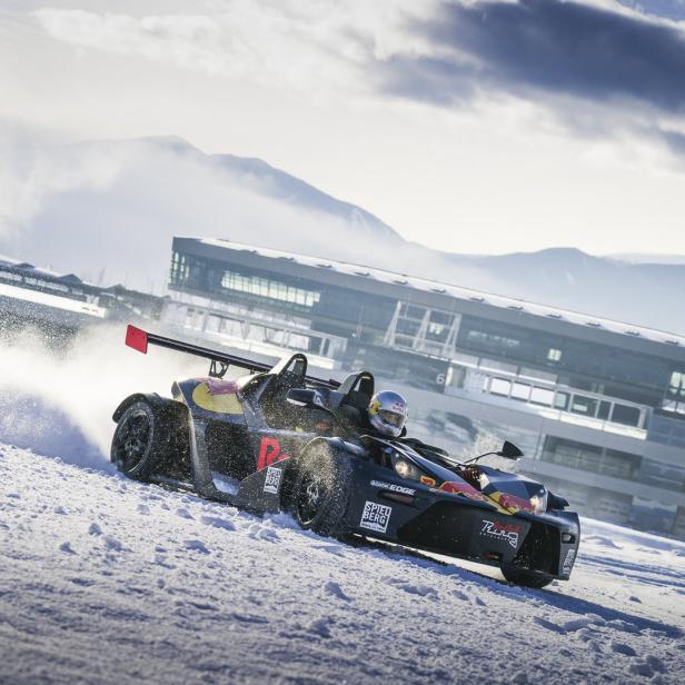 Ein KTM X-Bow fährt auf einer schneebedeckten Strecke, im Hintergrund ein Gebäude und Berge.