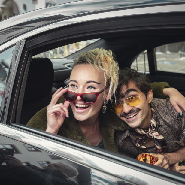 Eine lachende Frau mit Sonnenbrille und ein Mann mit gelber Brille sitzen in einem Auto.