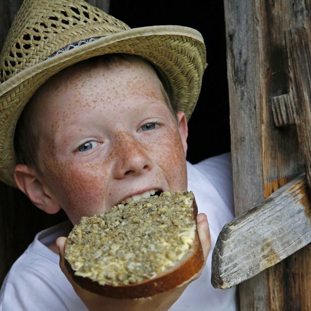 Ein Junge mit Sommersprossen und Strohhut isst ein belegtes Brot vor einer Holztür.