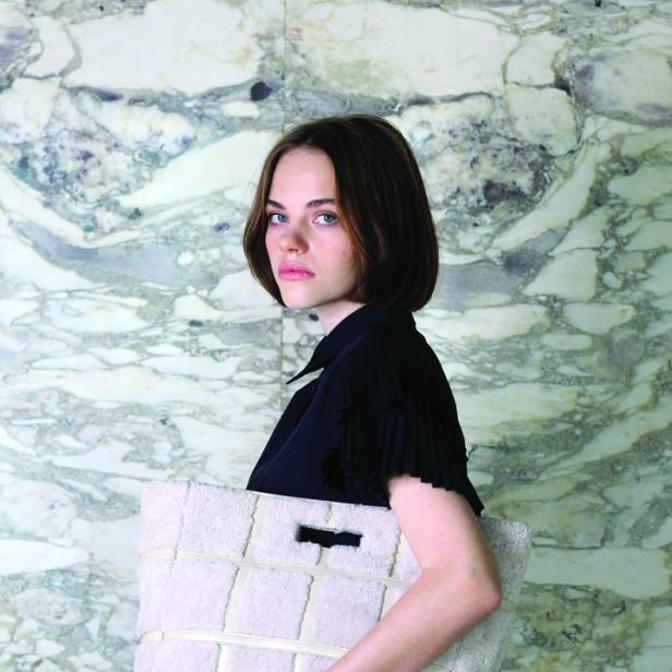 Eine Frau in einem schwarzen Kleid hält eine große, helle Tasche vor einem Marmorhintergrund.