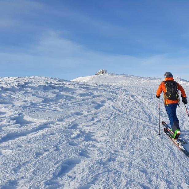 Ein Skitourengeher steigt auf einem verschneiten Hang in Richtung eines Gebäudes auf dem Gipfel.