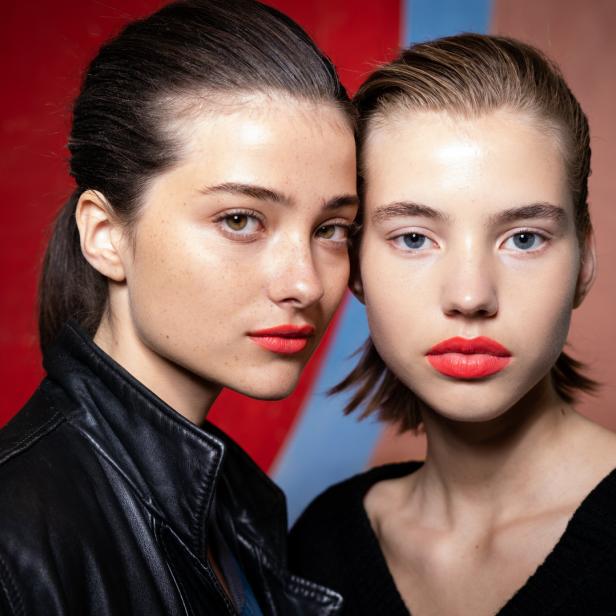Zwei junge Frauen mit leuchtend rotem Lippenstift posieren vor einem farbigen Hintergrund.
