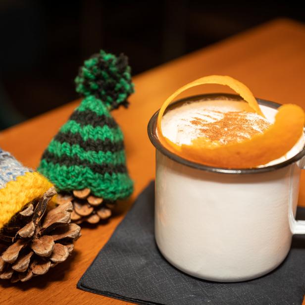 Ein Getränk mit Orangenschale in einer Tasse, neben zwei Tannenzapfen mit Miniaturmützen.