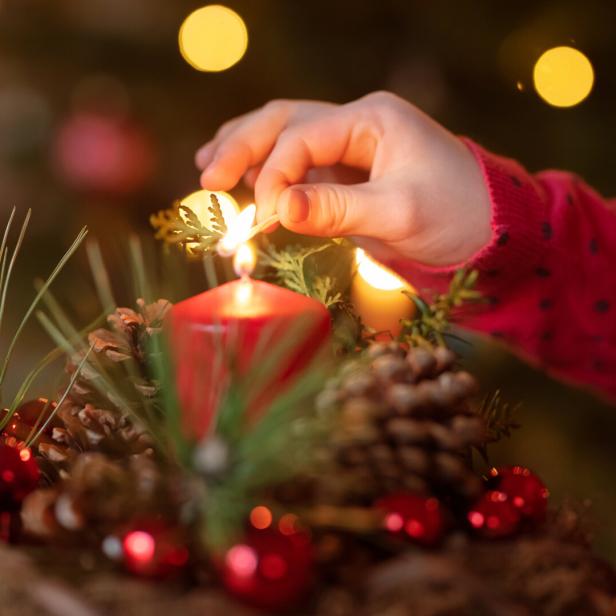 Eine Hand zündet eine rote Kerze auf einem weihnachtlich geschmückten Adventskranz mit Tannenzweigen und Zapfen an.