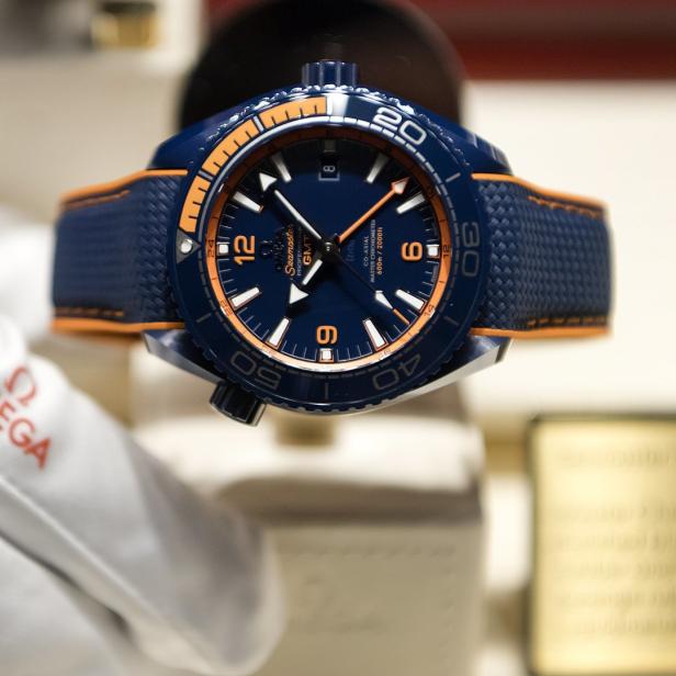 Eine blaue Omega Seamaster GMT Uhr mit orangefarbenen Akzenten wird von einem behandschuhten Hand präsentiert.