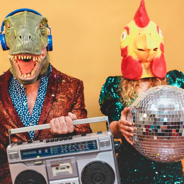 Eine Person mit Dinosaurier-Maske, Kopfhörern und Ghettoblaster neben einer Person mit Hühnermaske und Discokugel.
