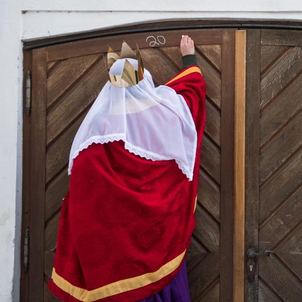Ein als Sternsinger verkleidetes Kind schreibt mit Kreide „20“ an eine Holztür.