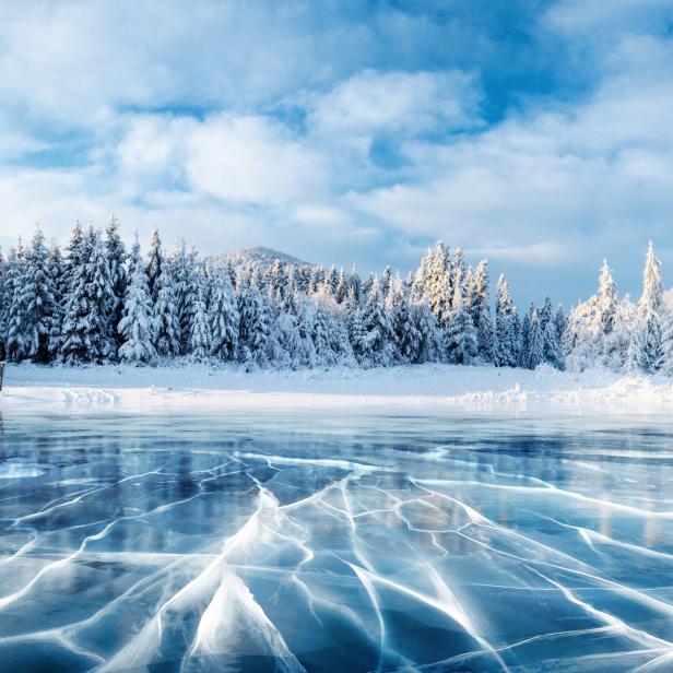 Ein zugefrorener See mit Rissen im Eis und schneebedeckten Bäumen im Hintergrund.