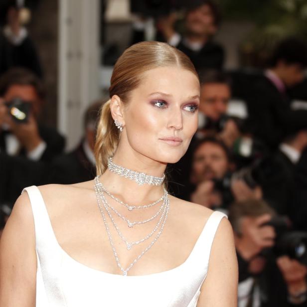 Eine blonde Frau mit weißem Kleid und auffälligem Schmuck posiert vor einer Menschenmenge.