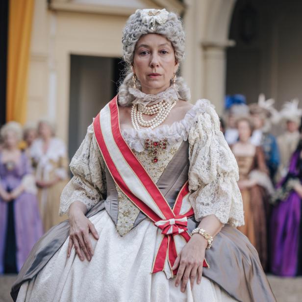 Eine Frau in historischer Kleidung mit Perlenkette und weiß-roter Schärpe vor einer Gruppe von Menschen.