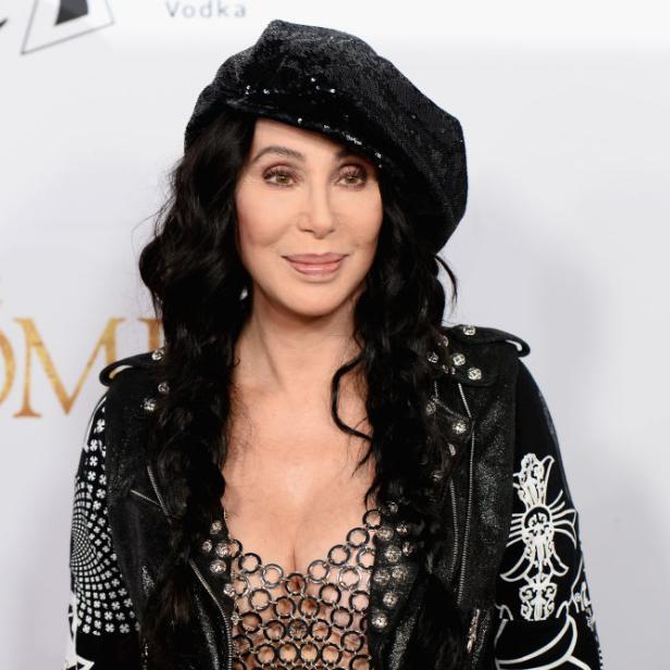 Cher posiert in einer schwarzen Jacke und einer passenden Mütze auf einem roten Teppich.
