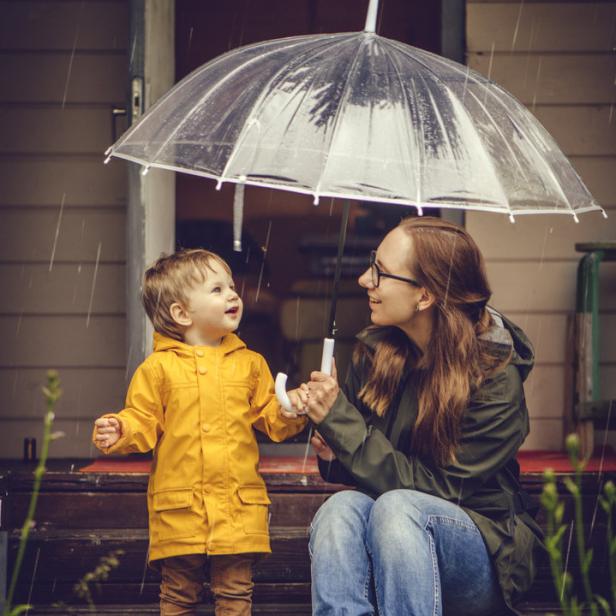 Eine Mutter und ihr Kind stehen mit einem Regenschirm im Regen vor einer Haustür.
