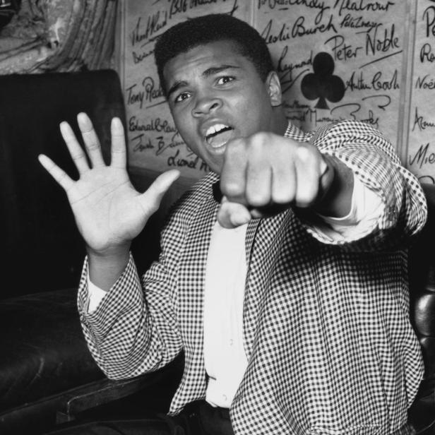 Muhammad Ali posiert mit erhobener Faust und offener Hand vor einer Wand mit Unterschriften.