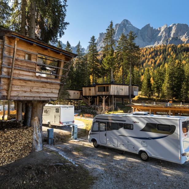 Ein Wohnmobil steht vor Baumhäusern in einer Waldlandschaft mit Bergen im Hintergrund.