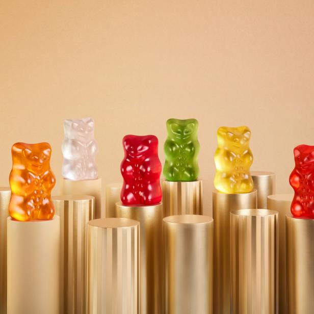 Mehrfarbige Gummibärchen stehen auf goldenen Säulen vor einem beigen Hintergrund.