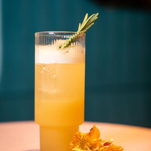 Ein erfrischender, orangefarbener Cocktail mit Rosmarinzweig und einer Blüte auf einem Tisch.
