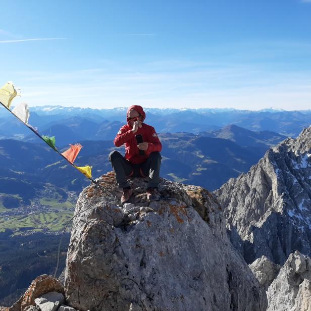Eine Person sitzt auf einem Berggipfel mit Gebetsfahnen und genießt die Aussicht.