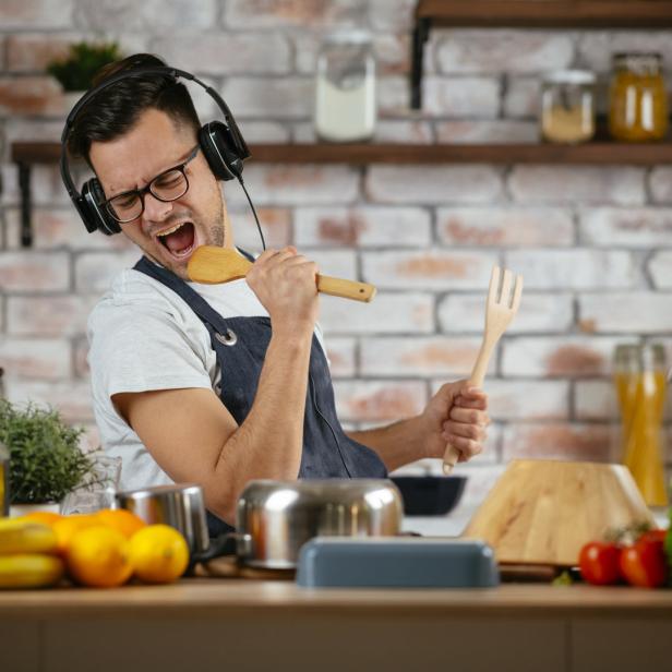 Ein Mann mit Kopfhörern singt mit einem Kochlöffel in der Küche.