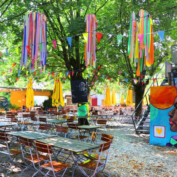Ein Biergarten mit Tischen, Stühlen, bunten Bändern und einem Wandgemälde.