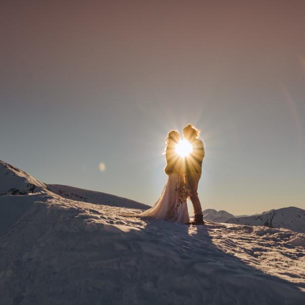 Ein Brautpaar umarmt sich auf einem schneebedeckten Berggipfel im Sonnenlicht.